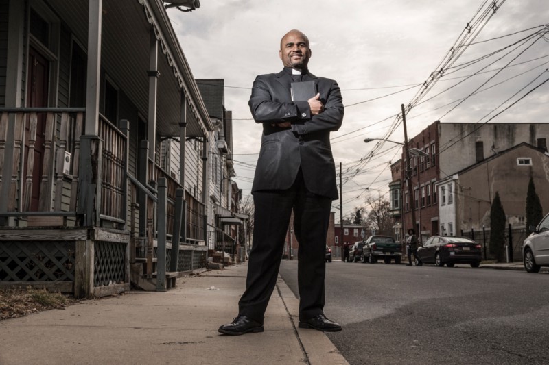 Armstrong stands on a street curb, dressed in all-black reverend attire. Surrounding him are run-down buildings. The sky is dark gray and power lines run overhead.