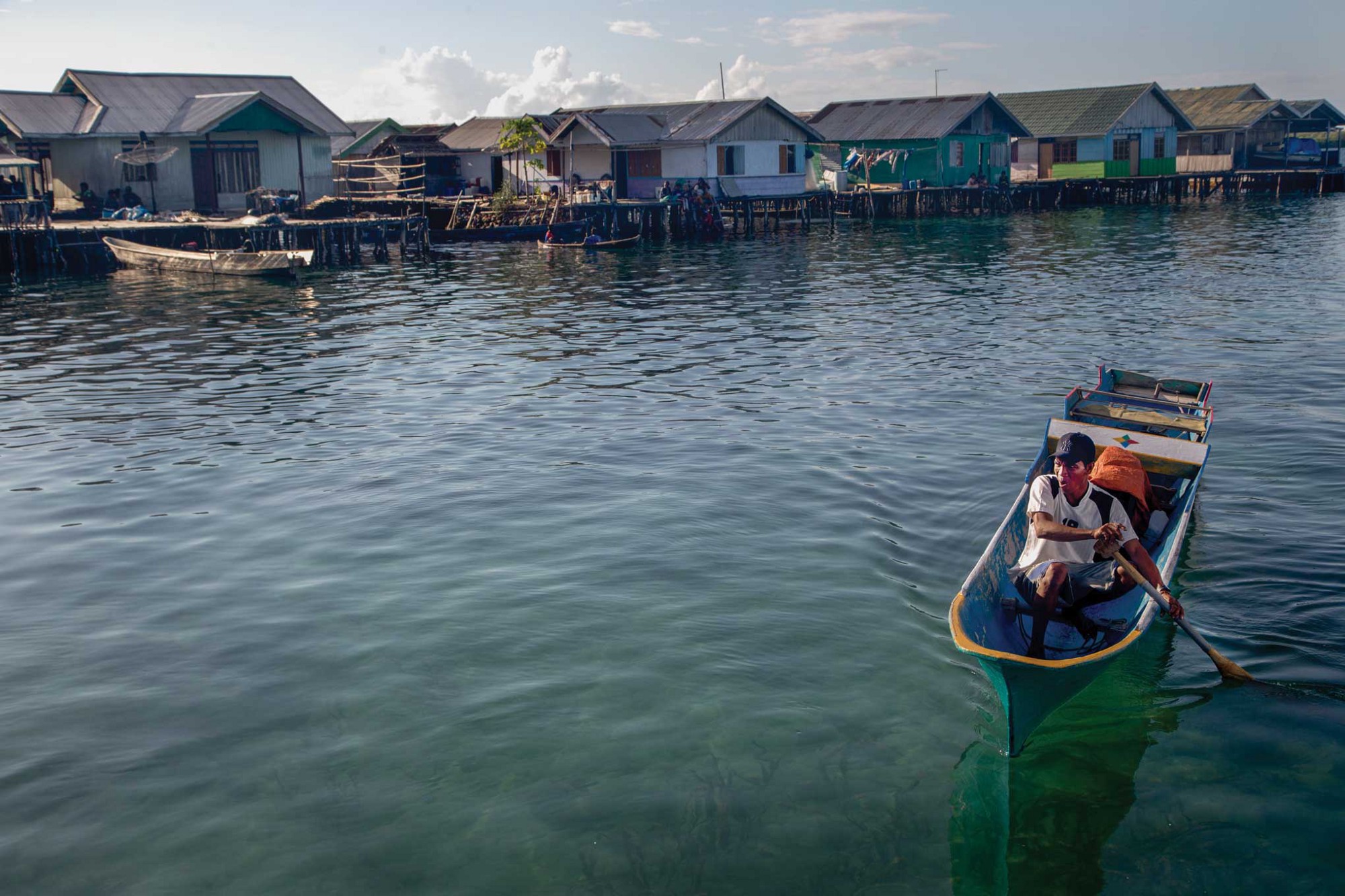 A man pilots a blue canoe just off the shore of a shanty town. He wears a baseball cap, some sort of jersey, and track shorts.