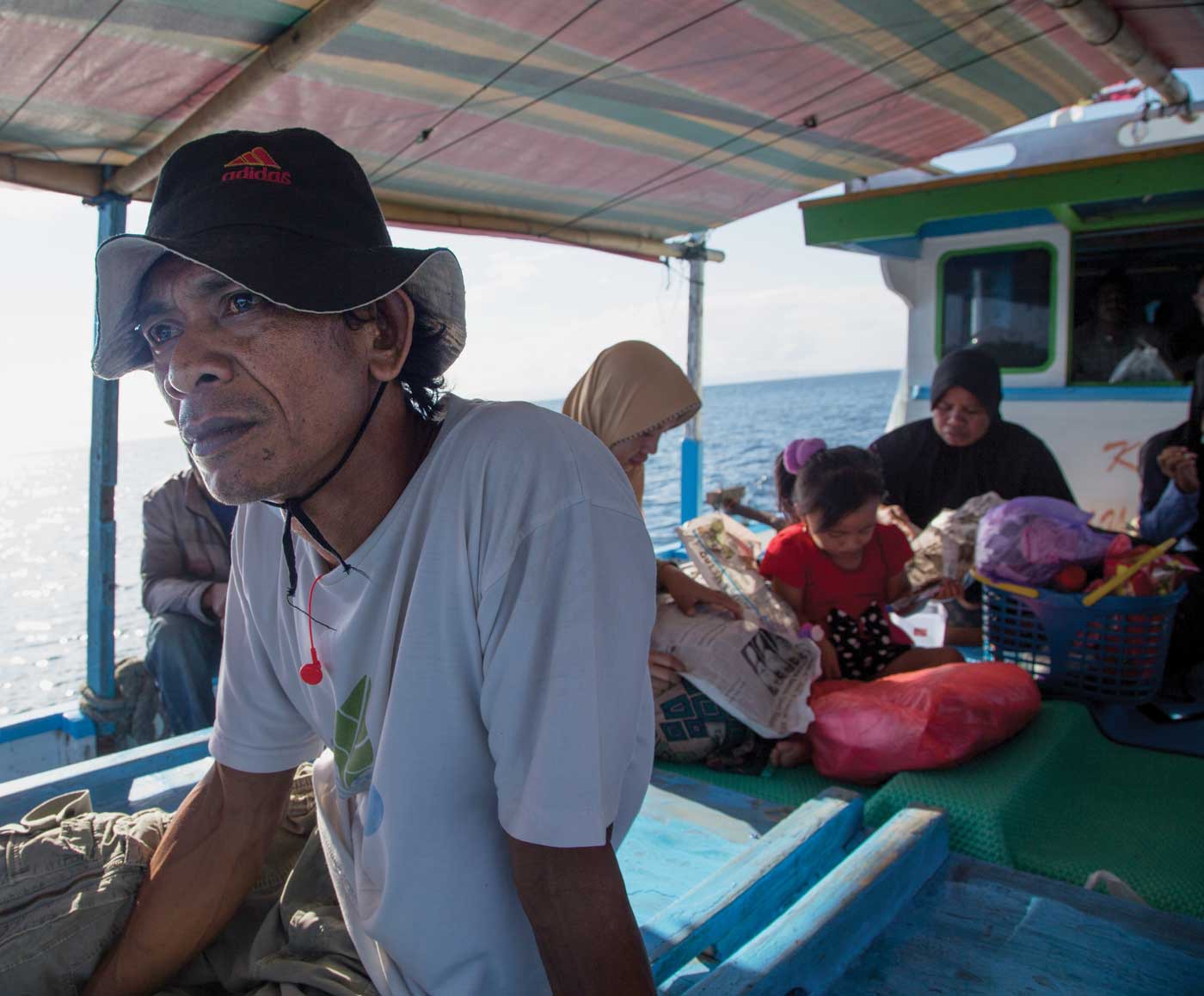 Sarly is shown on a boat, close-up, and looking to the left of the camera. He is wearing a fisherman's hat, a t-shirt, and cargo shorts. Behind him are two women and a child, all occupied with something.